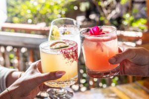 Three colorful cocktails raised for a toast on an outdoor patio.